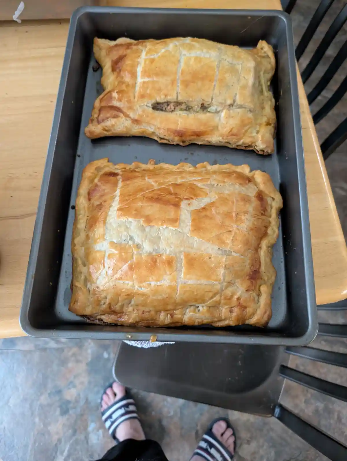 Two golden-brown, rectangular puff pastry pies sit in a metal baking tray on a wooden table. The top pie has a neat grid…