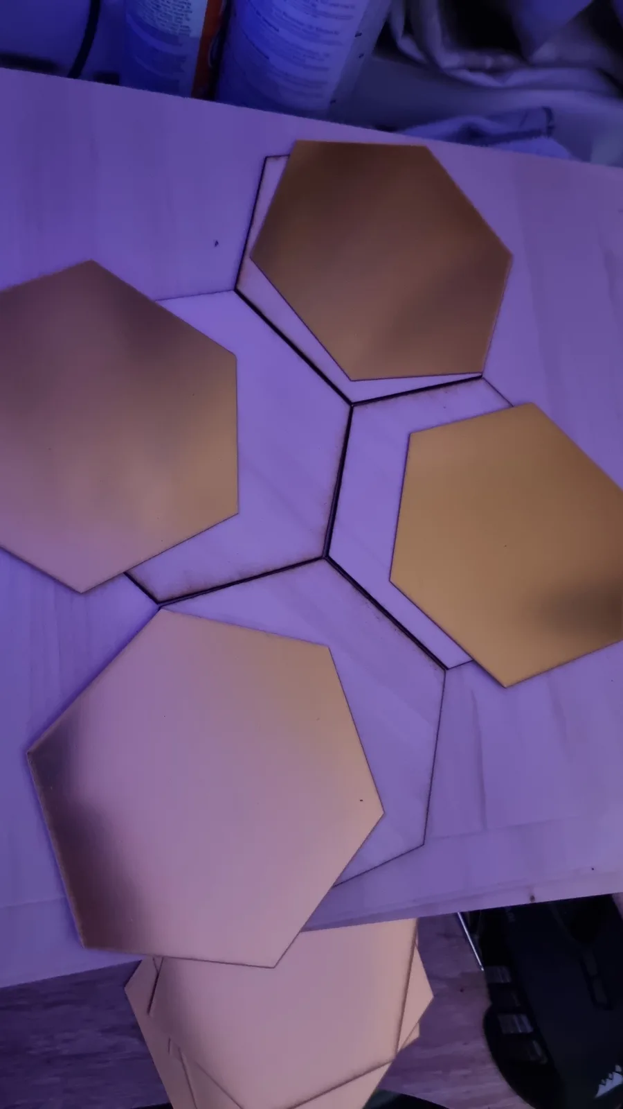 A close-up photo shows several gold-colored hexagonal mirror tiles arranged in a pattern on a white surface, under purple…