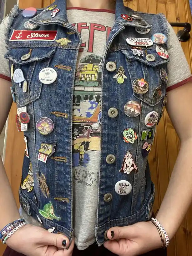 A person wears a heavily decorated denim vest covered in numerous colorful pins and badges, including one that reads…