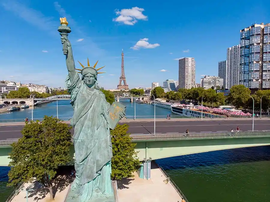 A large statue resembling the Statue of Liberty stands in a cityscape with the Eiffel Tower visible in the background…
