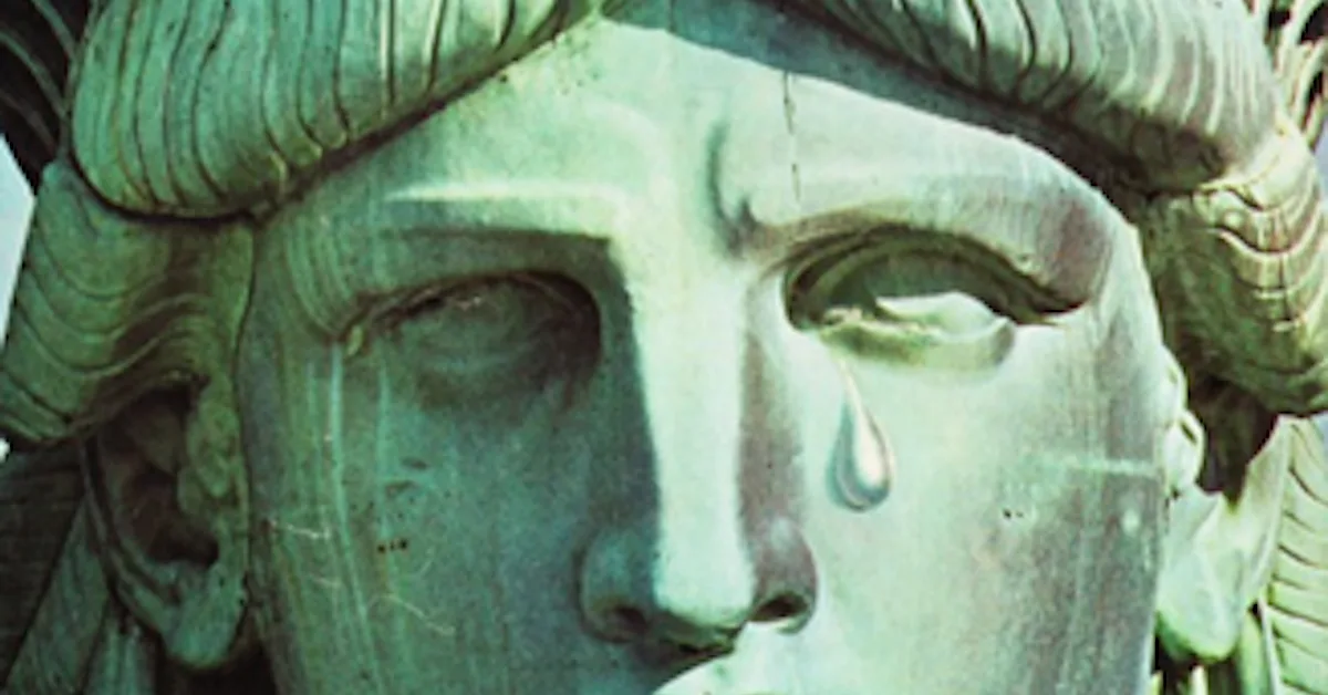 A close-up of a weathered, greenish statue of a person with one eye closed and a single teardrop falling from the other eye…