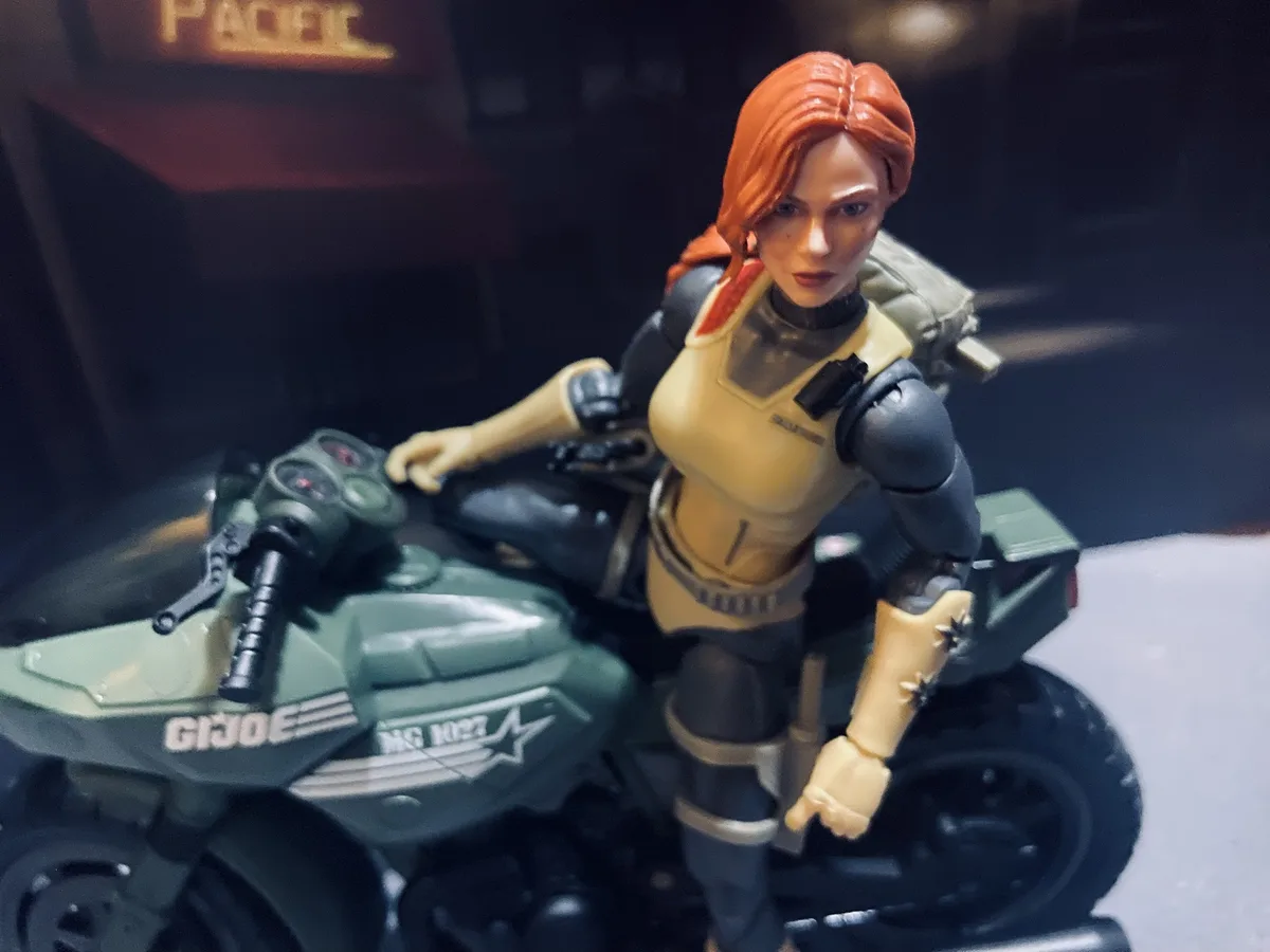 A G.I. Joe action figure with red hair, wearing a beige and black tactical outfit, is posed sitting on a green motorcycle…