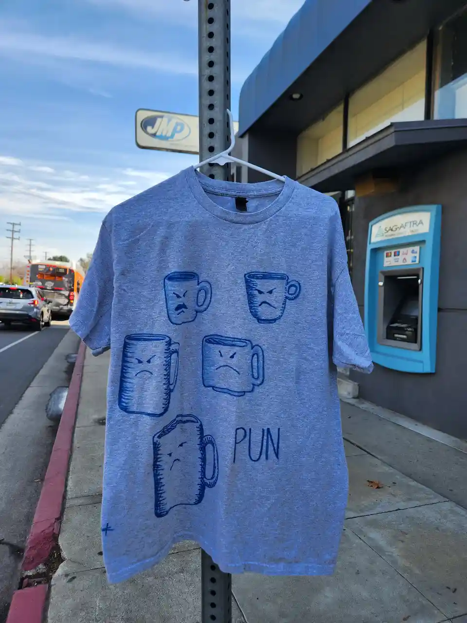 A blue t-shirt hangs on a white hanger from a metal pole on a sidewalk, featuring five cartoon coffee mugs with angry or…