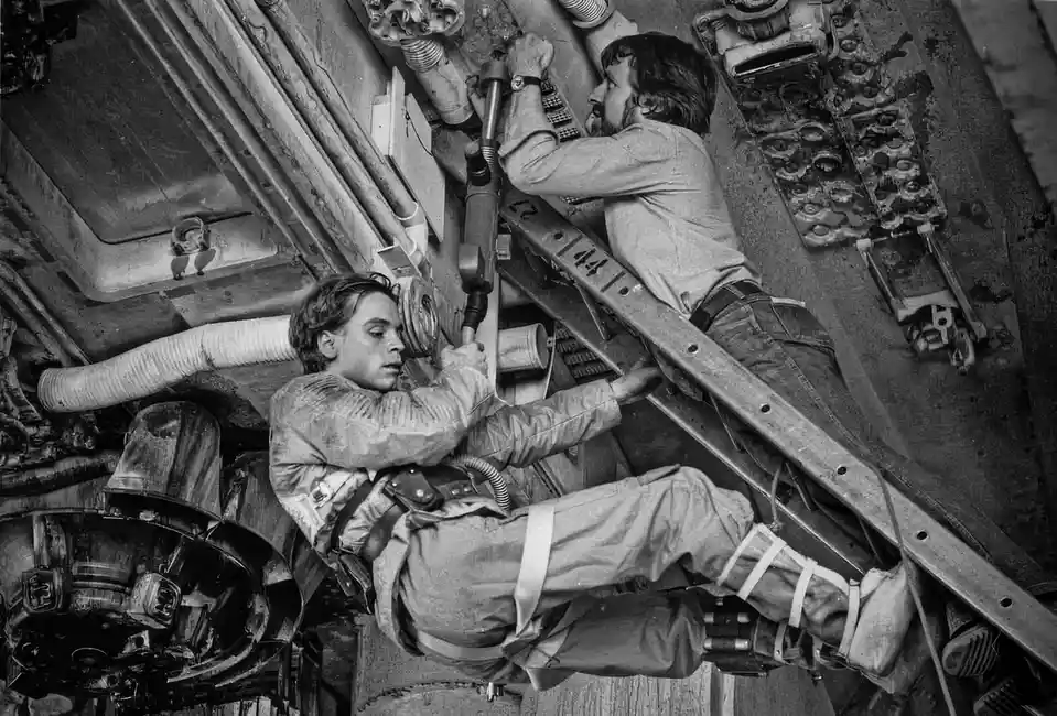 A black-and-white photo shows two men in a cramped, industrial setting, possibly inside a ship or machinery. One man is…