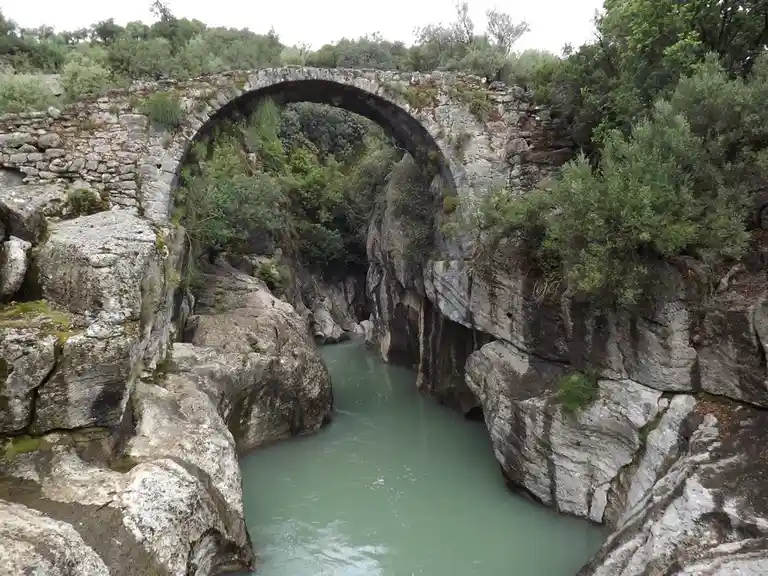 A weathered stone arch bridge spans a narrow canyon with greenish water flowing beneath it, surrounded by rocky cliffs and…