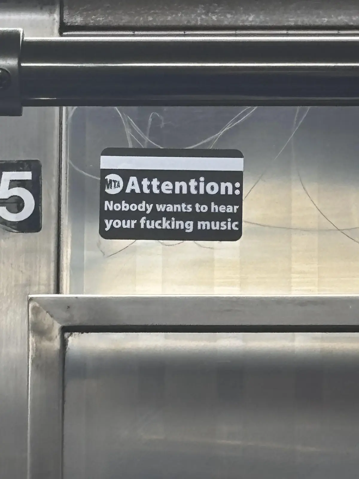 A black sticker with white text reading 'Attention: Nobody wants to hear your fucking music' is affixed to a glass panel of…