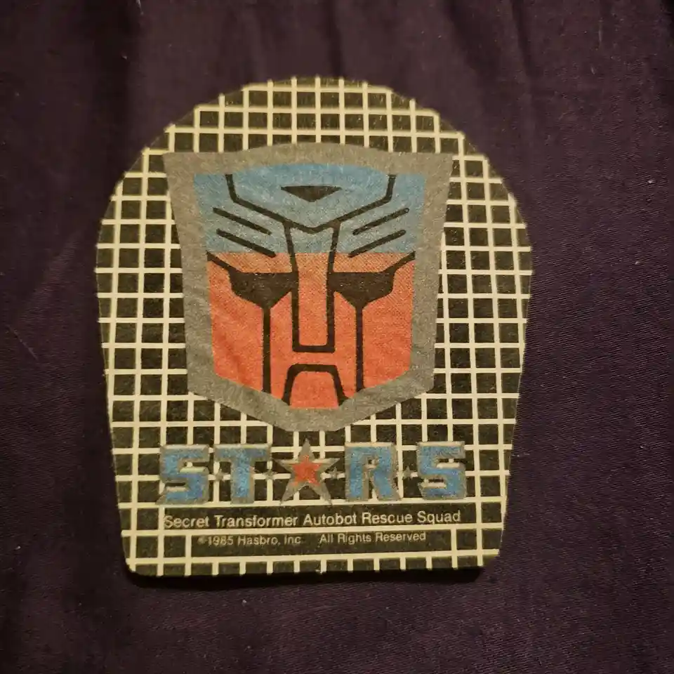 A close-up of a vintage Transformers STARS patch on a dark purple fabric, featuring the Autobot symbol in blue and red…