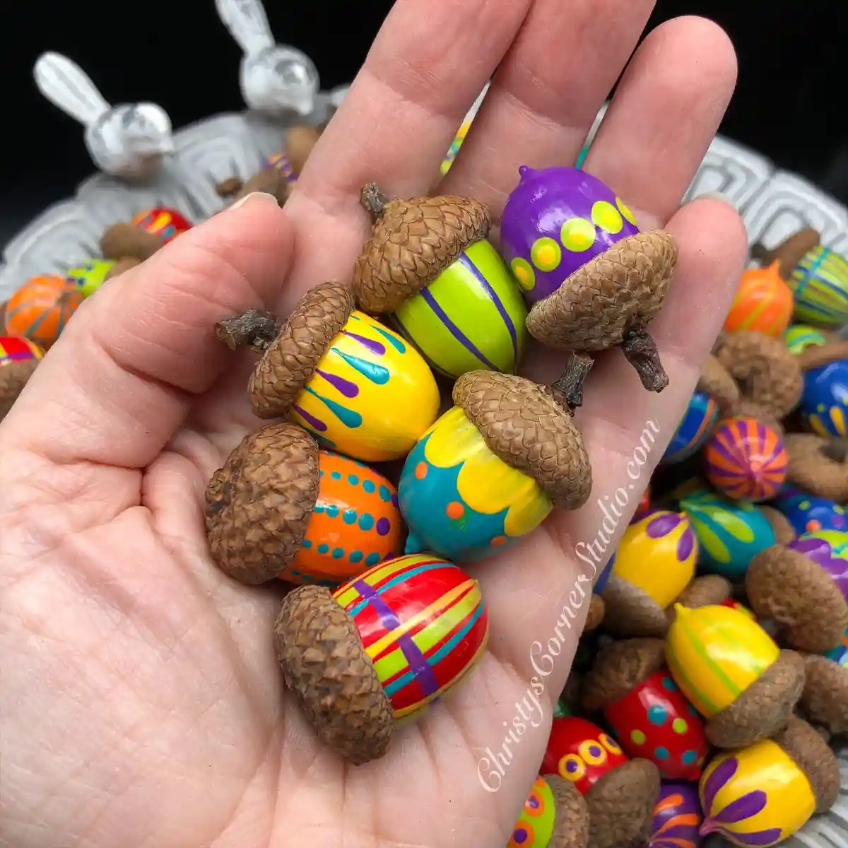 A hand holds several acorns that have been painted with bright, colorful patterns resembling Easter eggs, including stripes…