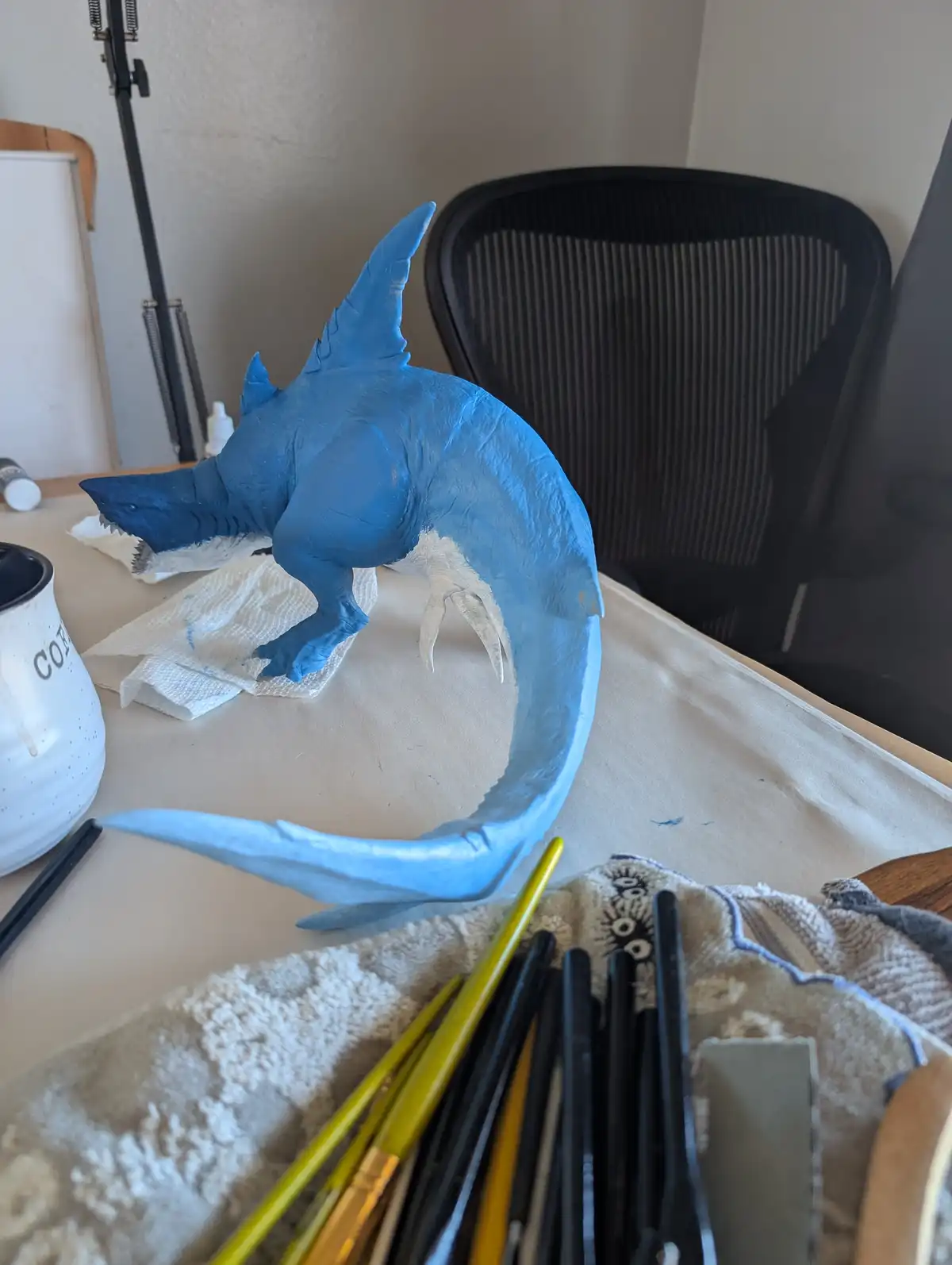 A blue-painted, shark-headed dragon sculpture sits on a desk surrounded by paintbrushes, paper towels, and a mug, with the…