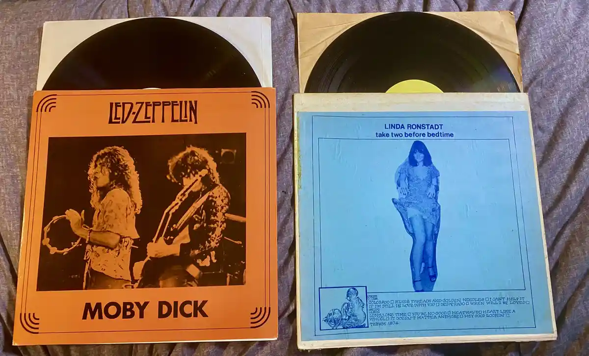 Two vinyl records are shown side-by-side on a gray fabric surface. The left record has an orange cover with 'LED-ZEPPELIN'…
