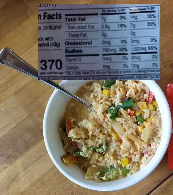 A white bowl of instant ramen noodles mixed with vegetables sits on a wooden table, with a metal spoon resting inside. A…