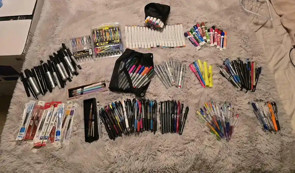 An overhead shot displays a large, organized collection of writing instruments—pens, markers, and pencils—arranged in neat…