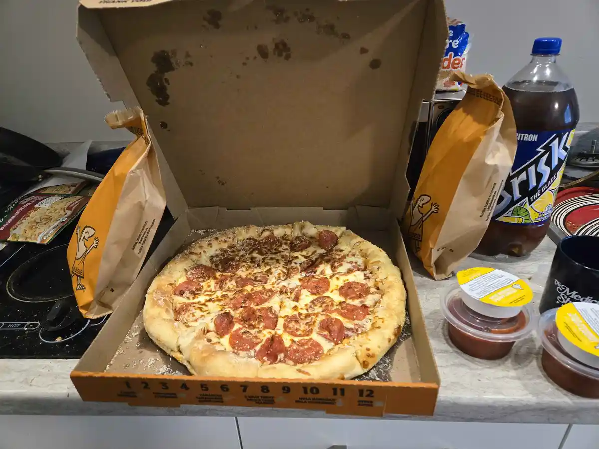 A partially eaten pepperoni pizza sits in an open cardboard box on a kitchen counter, surrounded by snack bags, a bottle of…
