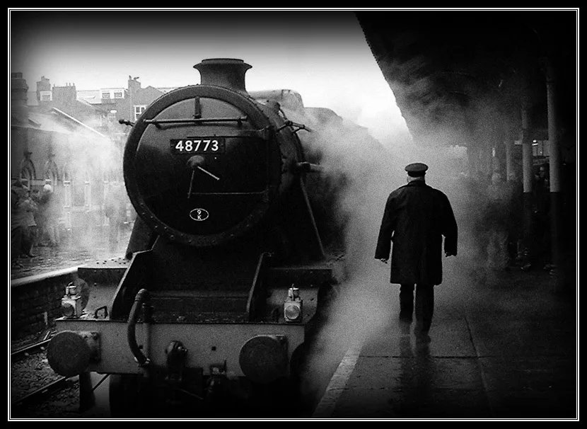 A black-and-white photo shows a steam locomotive, numbered 48773, at a station platform with steam billowing. A man in a…