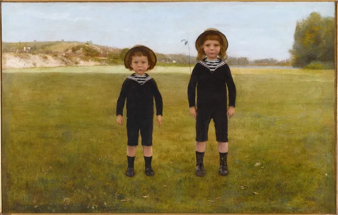 Two young boys dressed in identical black sailor suits with white-trimmed collars and straw hats stand side-by-side in a…
