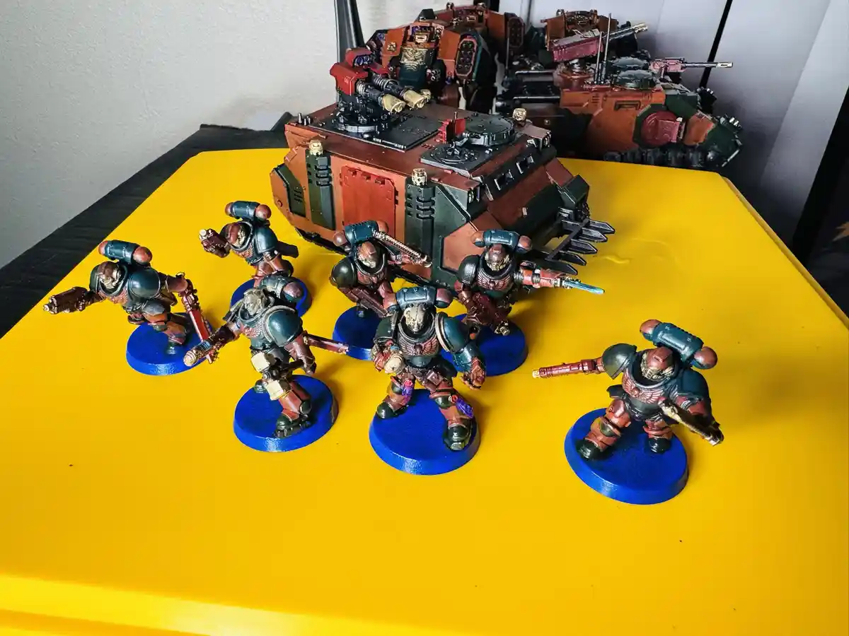 A photo of six painted miniature warhammer 40k space marines and a vehicle on a bright yellow surface, with overlaid text…
