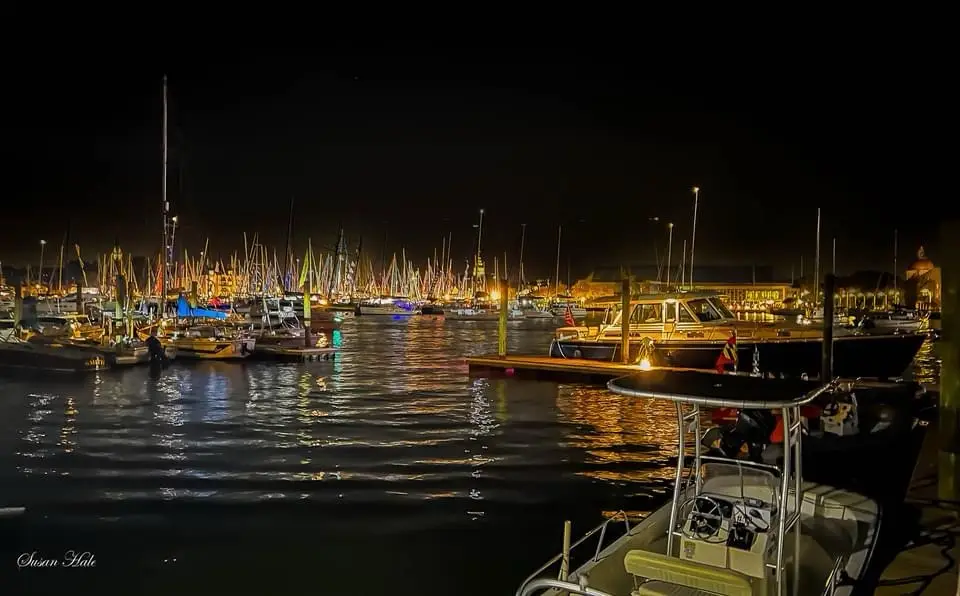 A nighttime photograph of a marina filled with numerous boats and yachts, their lights reflecting on the water. The…