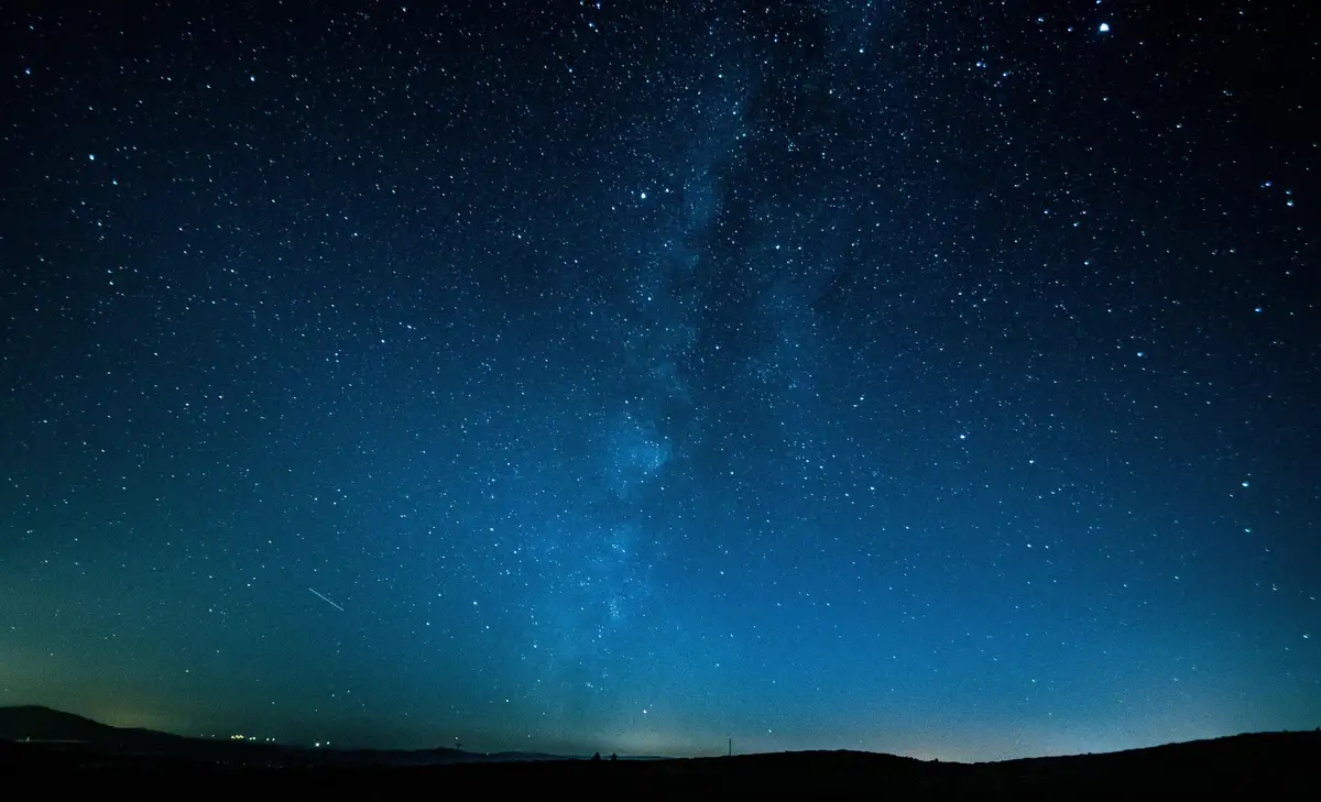 A wide-angle night sky photograph shows a dense field of stars with the Milky Way galaxy prominently visible as a bright…