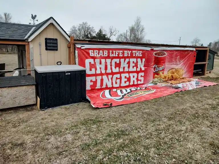 A large red Cane's Chicken Fingers advertisement banner is draped over a wooden structure resembling a chicken coop in a…