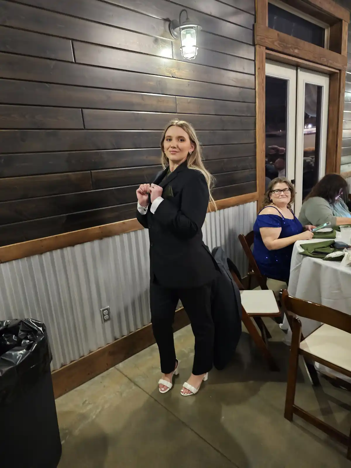 A young woman in a black tuxedo suit with white heels poses confidently indoors, adjusting her jacket. Behind her, a woman…