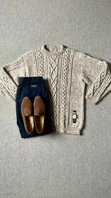 A flat-lay image displays a beige cable-knit sweater, navy blue chinos, brown loafers, and a wristwatch arranged neatly on a…