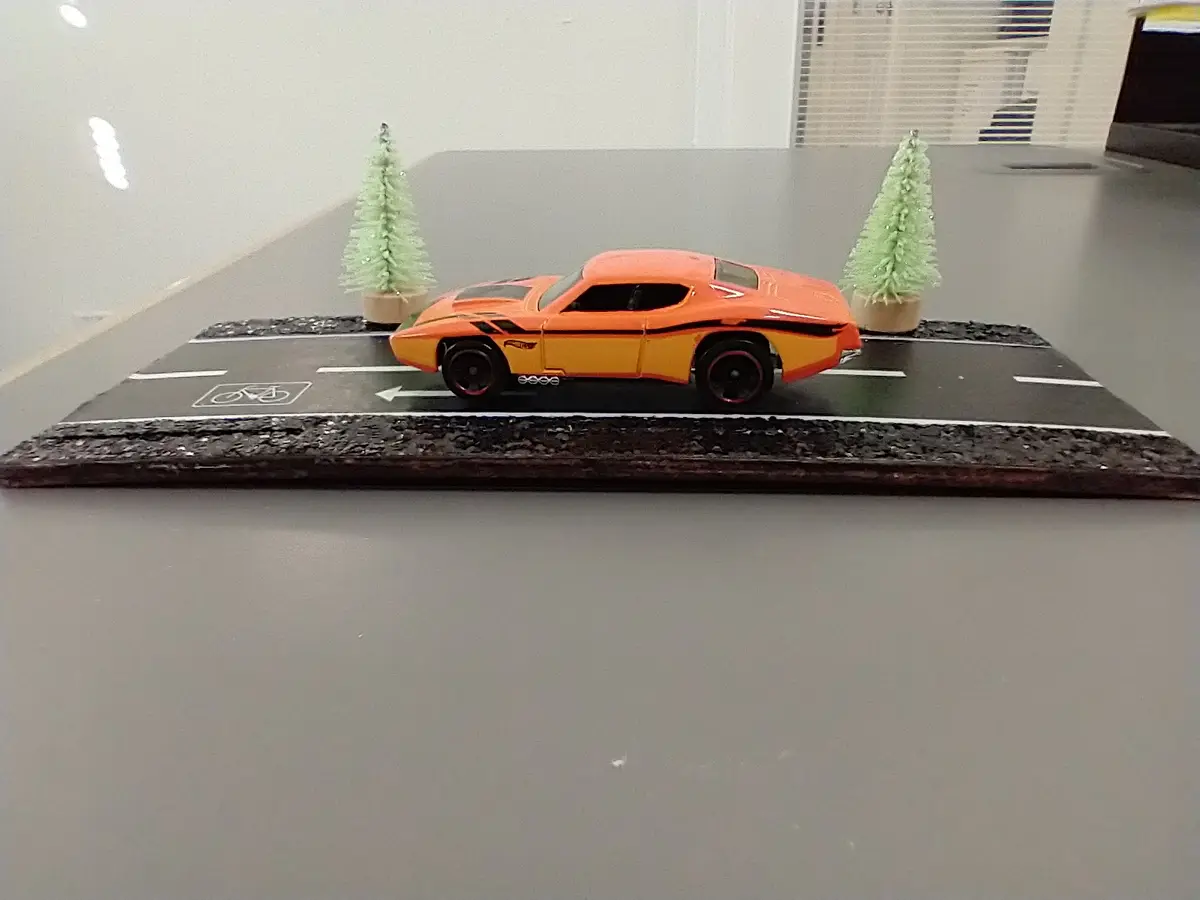 A small orange and yellow Hot Wheels-style toy car is positioned on a miniature road with white lane markings, flanked by…