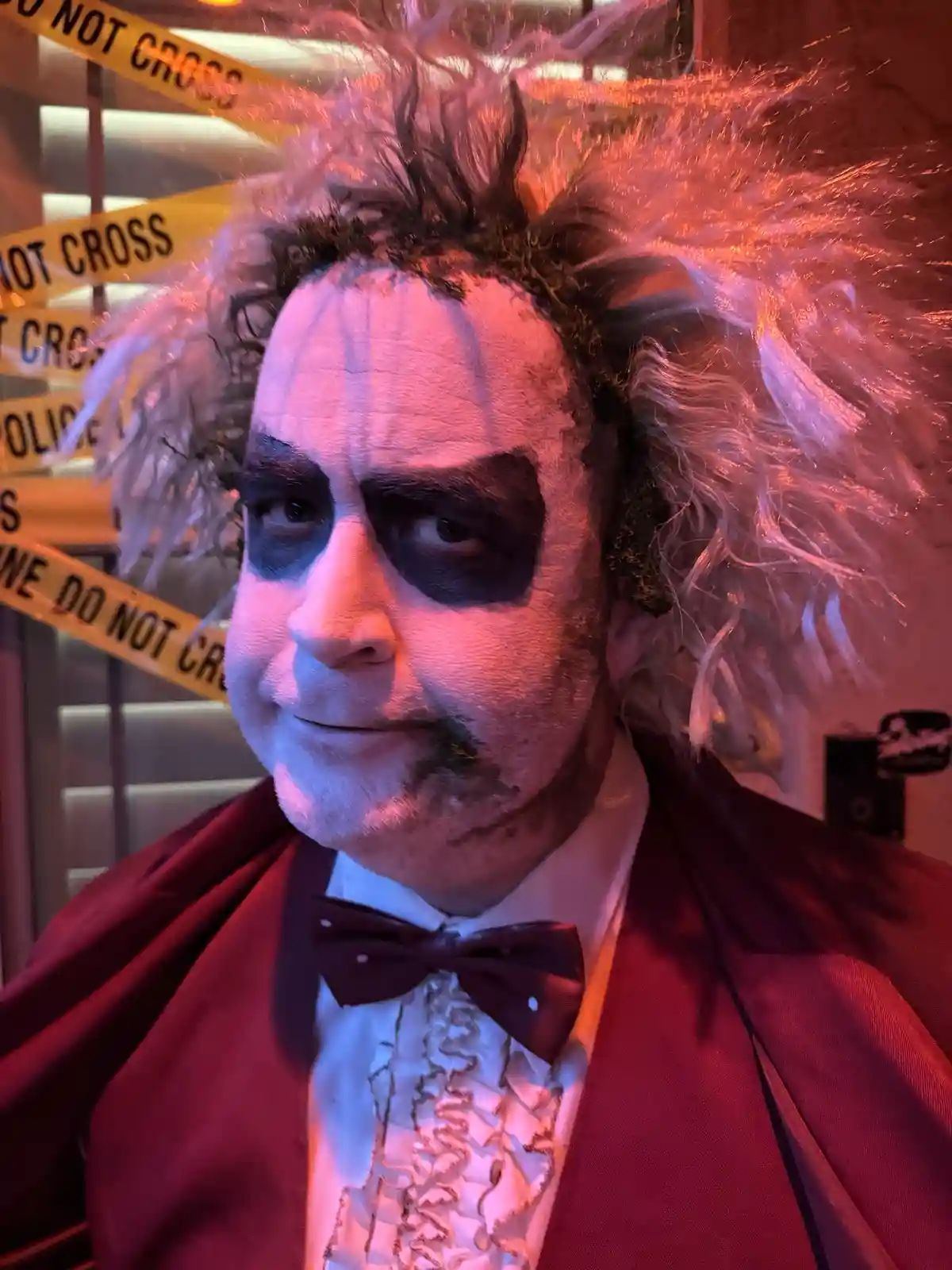 A person dressed as Beetlejuice with white face paint, black eye makeup, and wild hair, wearing a red cape and bow tie…