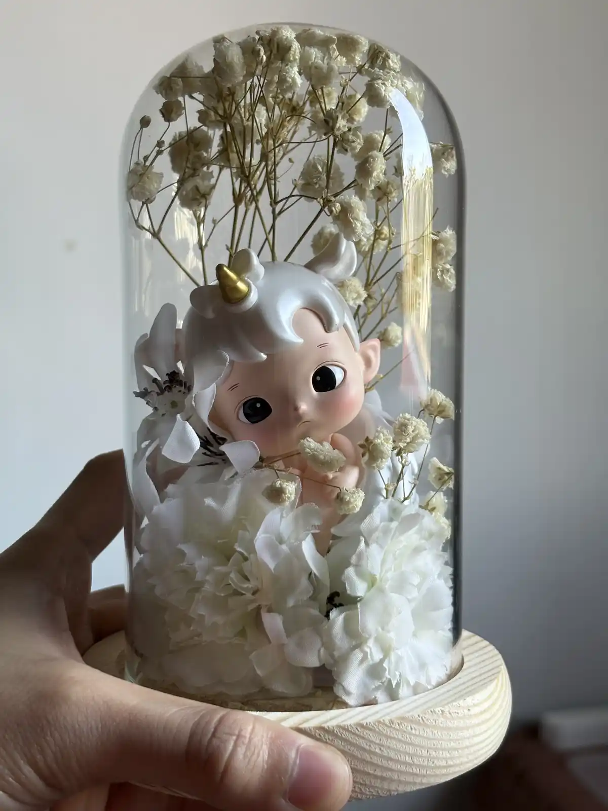 A hand holds a glass cloche containing a small figurine of a baby with white hair, a golden unicorn horn, and large black…