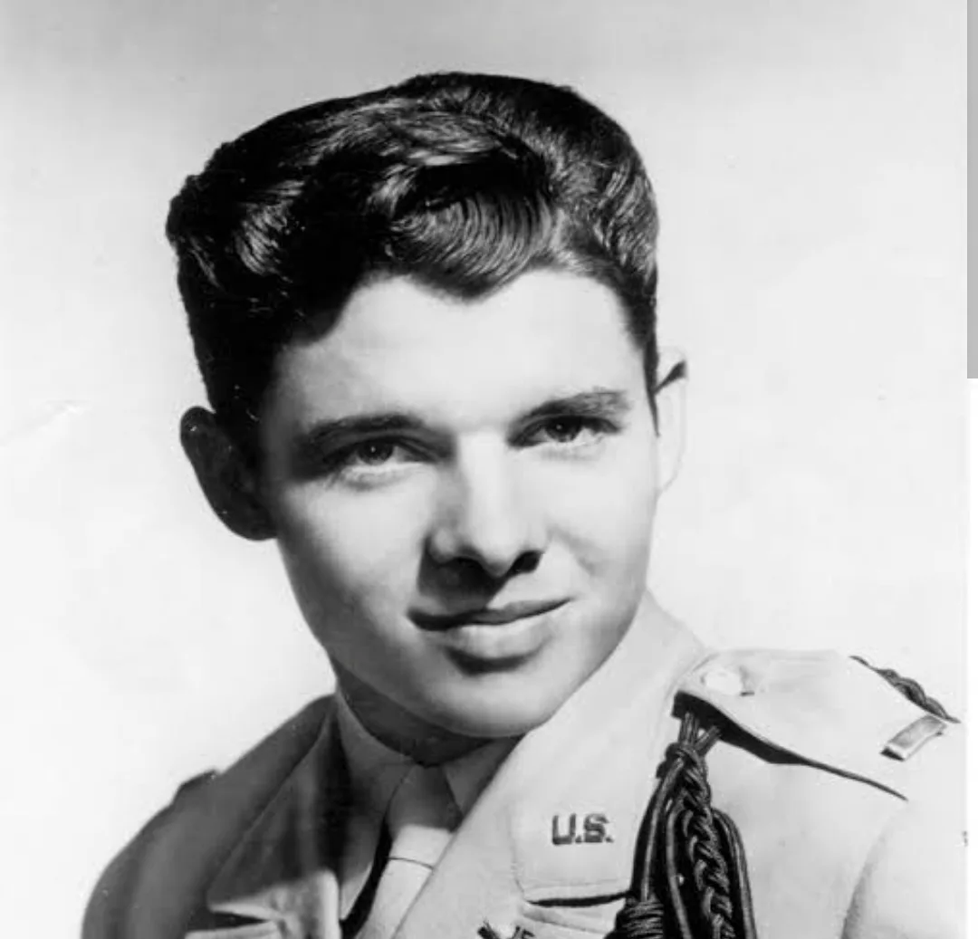 A black-and-white portrait of a young man in a U.S. military uniform with braided cords on his shoulder, smiling directly at…