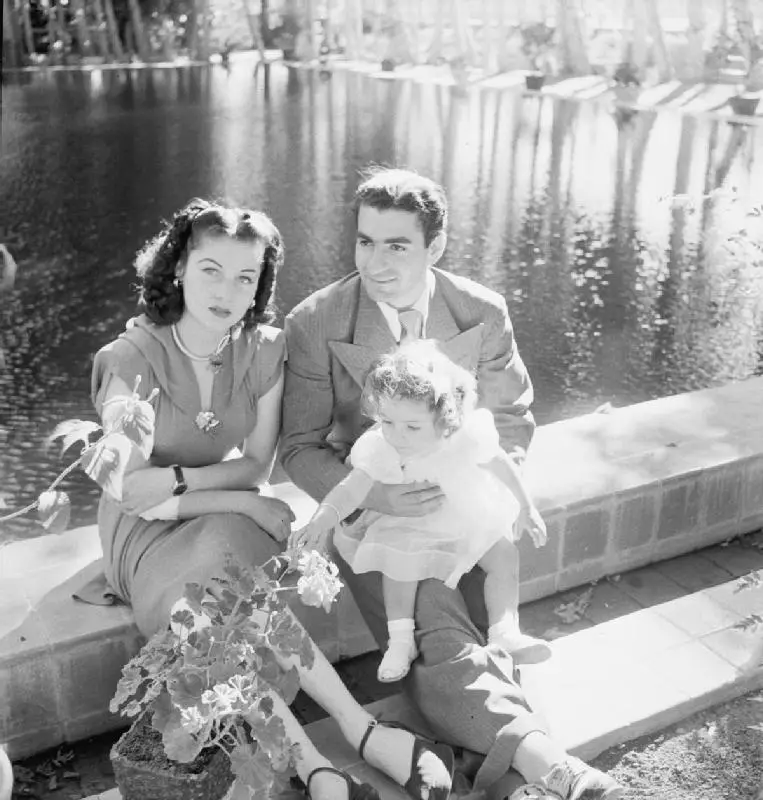 A vintage black-and-white photo shows a man, woman, and small child posing by a pond. The woman looks away with a…