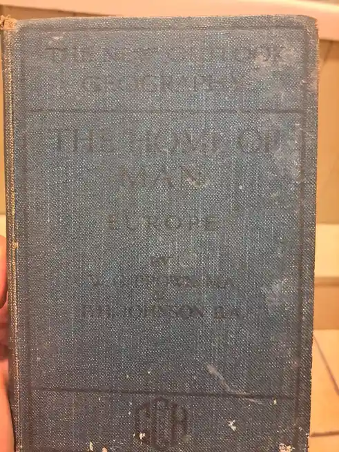 A close-up of a worn, dark blue cloth-bound book cover with faded embossed text. The visible title reads 'THE HOME OF MAN…