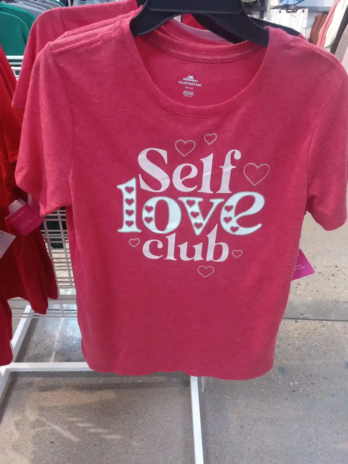 A red t-shirt hanging on a black hanger in a retail store displays the phrase 'Self love club' in large white letters…