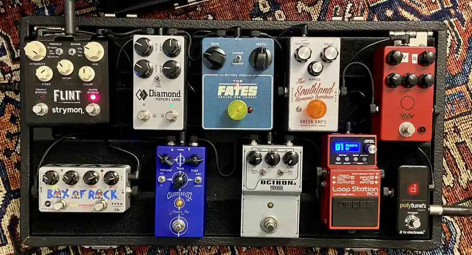 A top-down photo of a black pedalboard holding nine different guitar effects pedals, each with knobs and switches, connected…