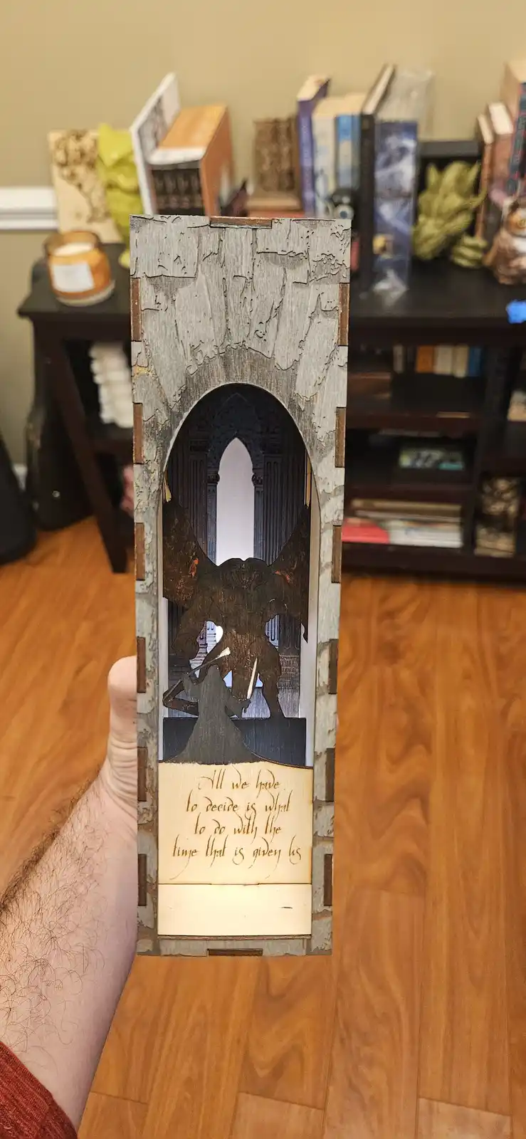 A hand holds a vertically oriented, intricately laser-cut wooden diorama depicting a dramatic scene: a small figure in a…