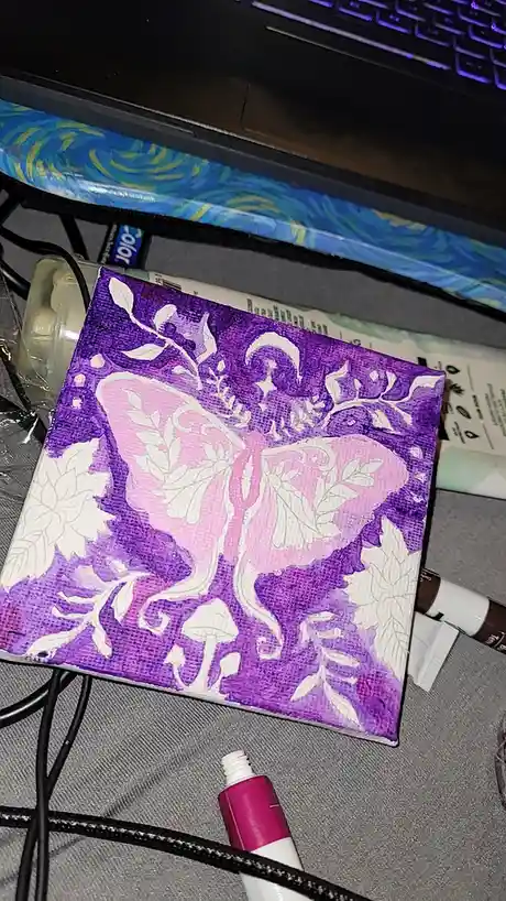 A small square canvas painting with a purple background and a large pink butterfly outlined in white, surrounded by white…