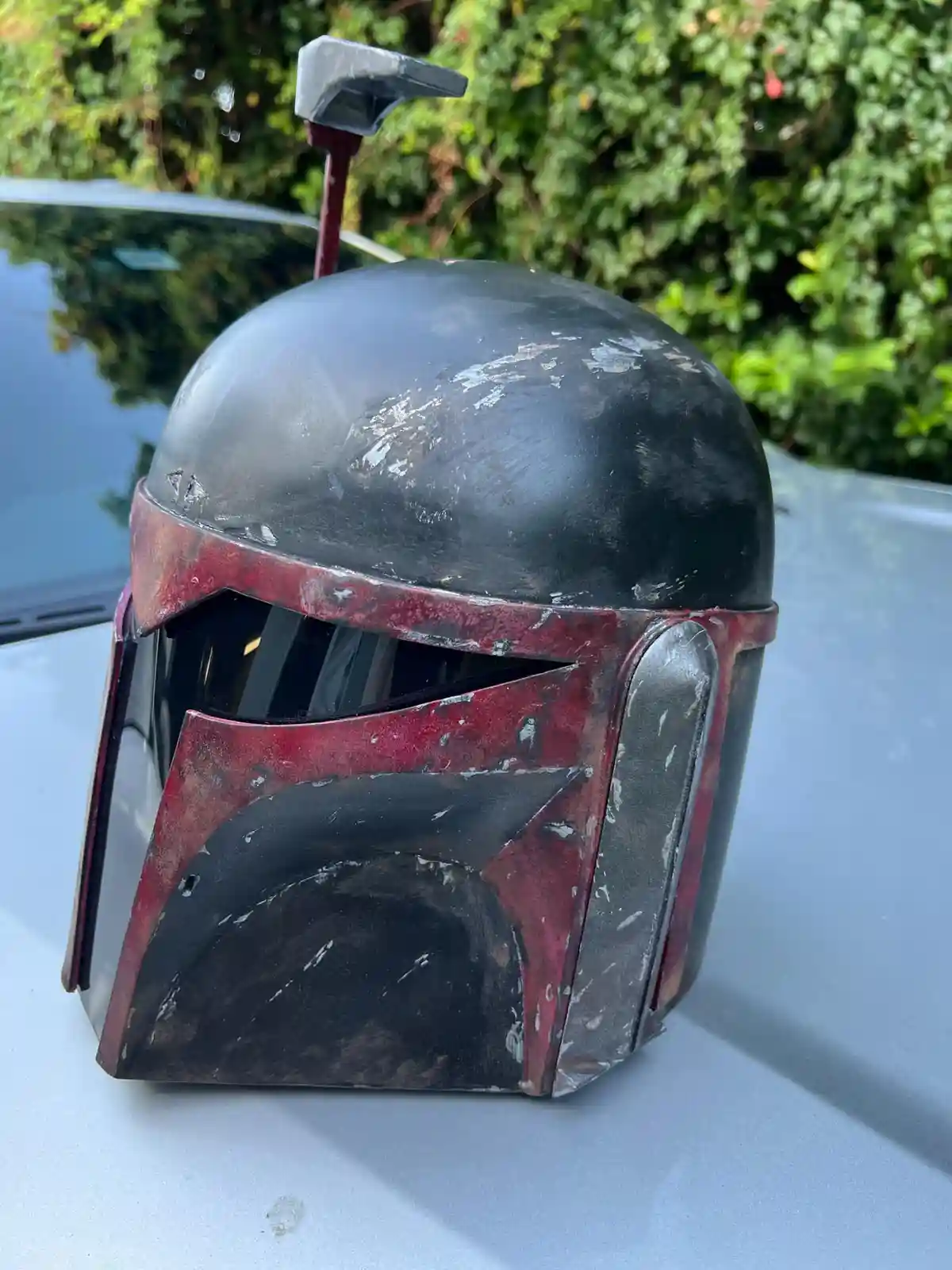 A weathered, black and red Boba Fett helmet from Star Wars sits on the hood of a car, with a small hammer stuck into its…