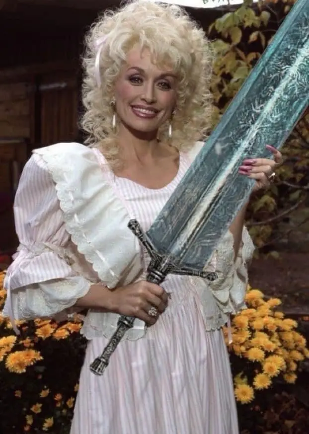 A woman with voluminous blonde curls and a pink and white striped dress holds a large ornate sword. She smiles at the camera…