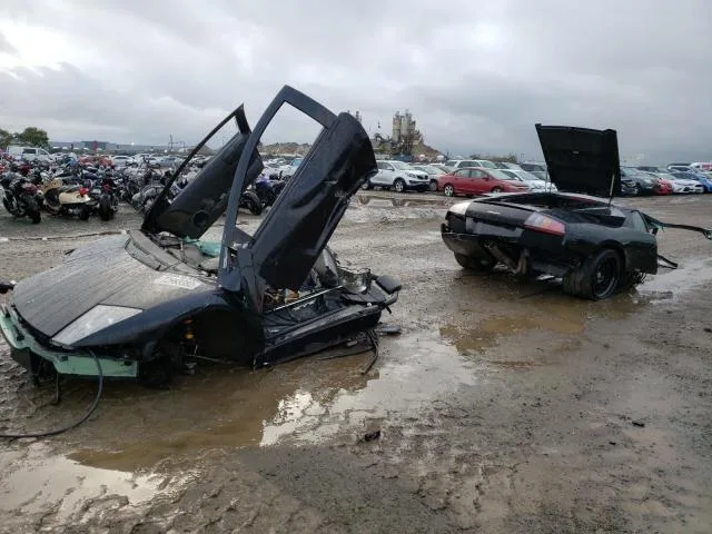 Two heavily damaged black Lamborghini Diablo sports cars sit in a muddy junkyard, one with its doors open and the other with…