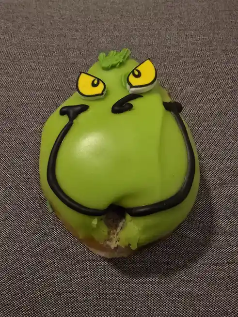 A close-up of a round, green-frosted donut decorated to resemble the Grinch, featuring yellow eyes with black outlines, a…