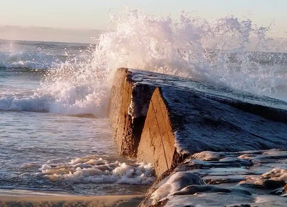 A large, weathered concrete sea wall or breakwater stands firm as ocean waves crash violently against it, sending a massive…