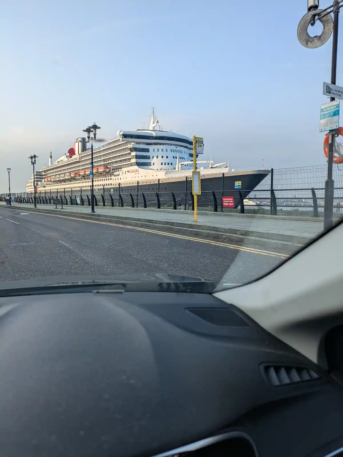 A photo taken from inside a car shows a large Cunard cruise ship docked at Liverpool Waters, viewed through a roadside…