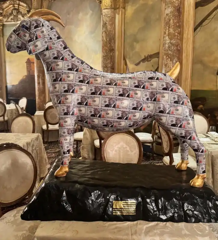 A large, ornate statue of a goat stands on a black pedestal in a luxurious dining room, its entire body covered in printed…