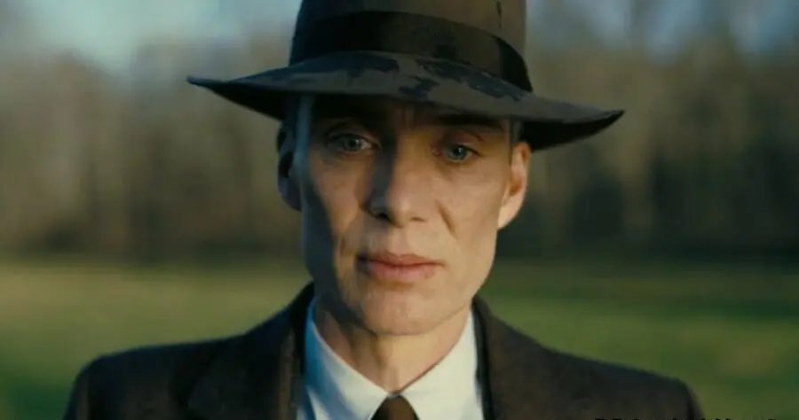 A man in a dark suit, white shirt, and brown tie stands outdoors in a field, wearing a dark fedora hat. His expression is…
