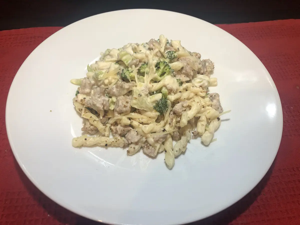 A white plate holds a creamy pasta dish with spiral noodles, chopped broccoli florets, and crumbled sausage, all coated in a…