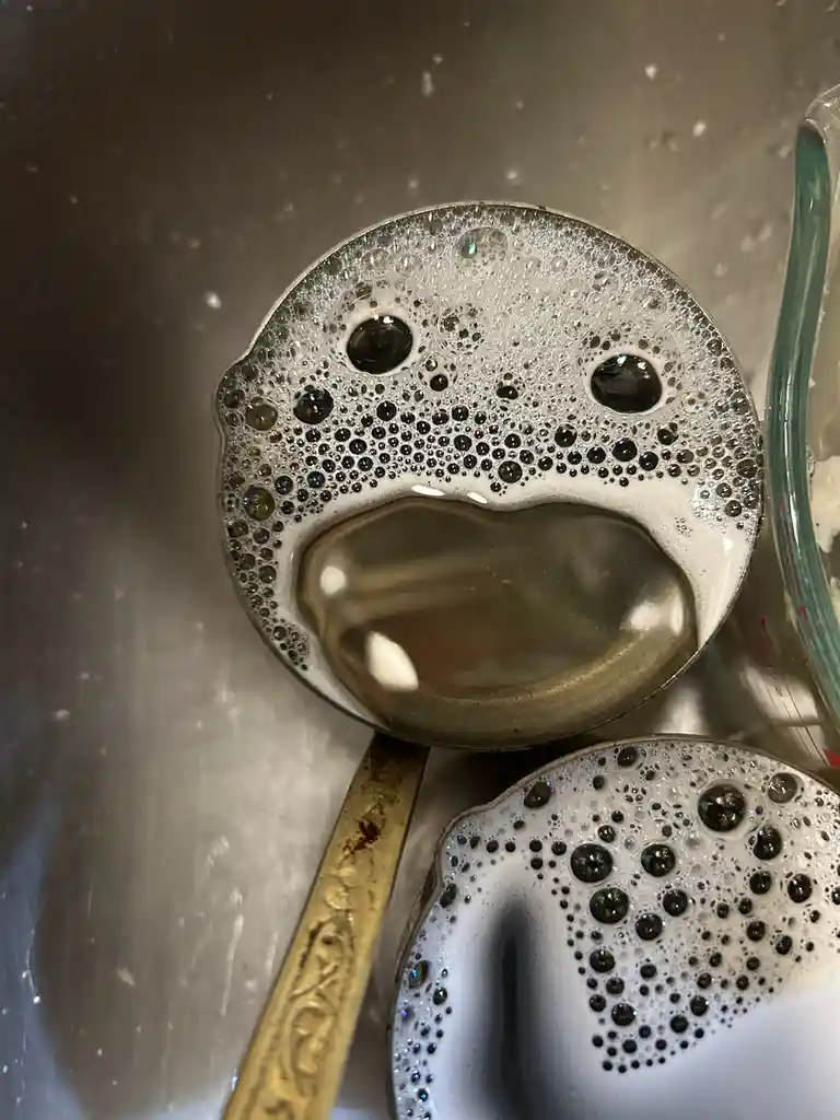A close-up photo shows two metal spoons in a stainless steel sink, covered in soapy bubbles that form a sad, crying face on…