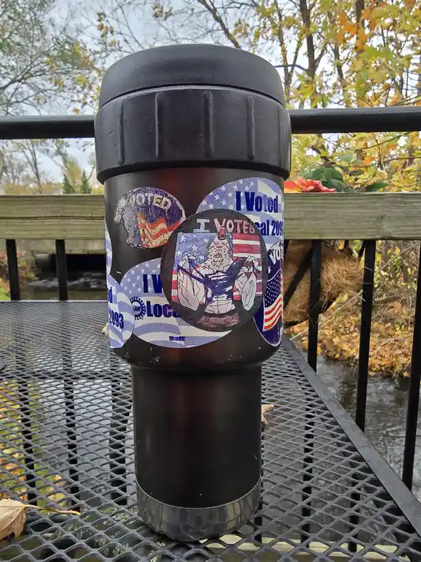 A black reusable travel mug sits on a metal mesh table outdoors, covered in multiple worn "I Voted" stickers from various…
