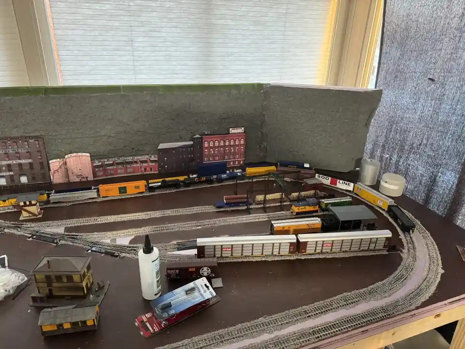 A detailed model train layout with multiple tracks, miniature buildings, and freight cars sits on a wooden table. A bottle…