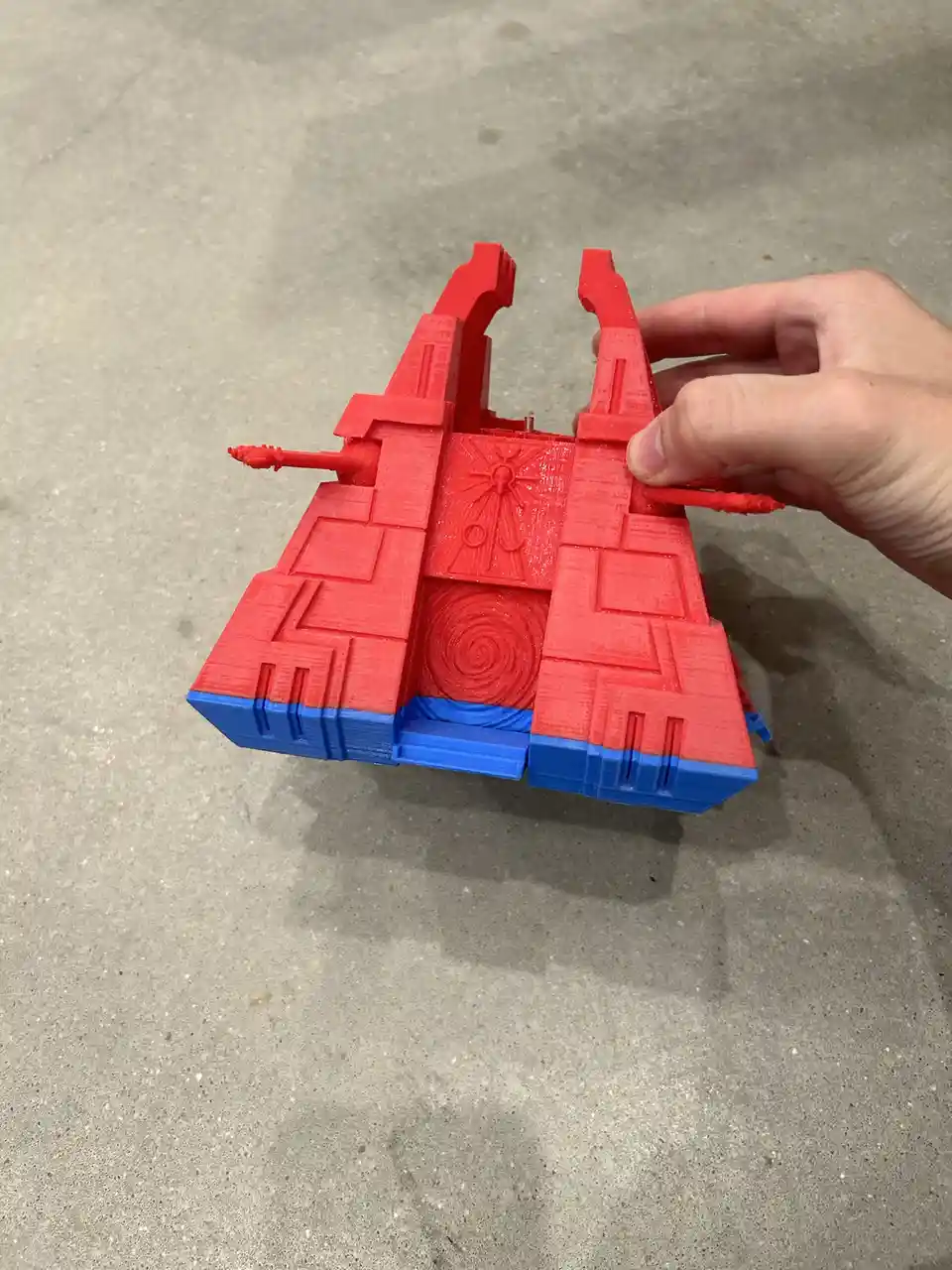 A hand holds a red and blue 3d-printed object resembling a stylized, angular Spider-Man suit or costume piece, with visible…