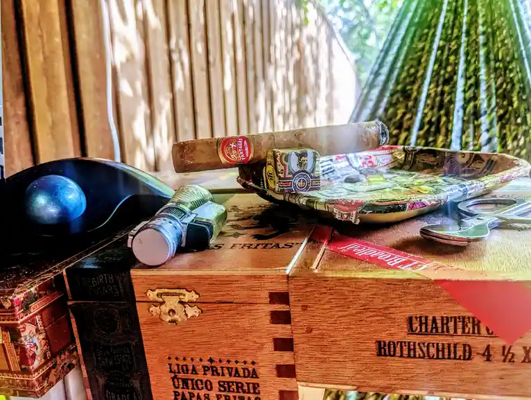 A lit cigar rests on a decorative ashtray atop a wooden cigar box labeled 'Liga Privada Único Serie' and 'Rothschild 4 ½ X'…
