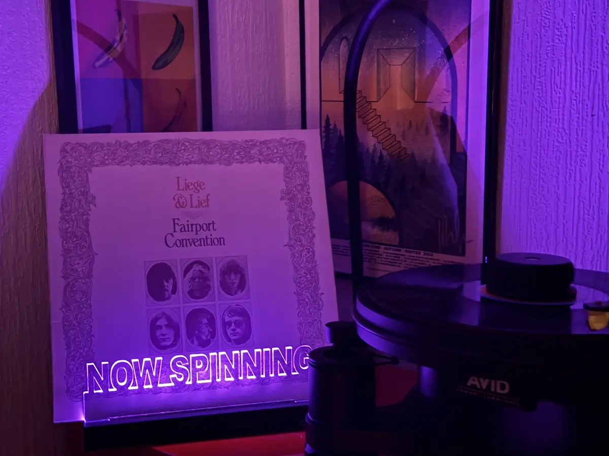 A vinyl record player with an 'AVID' brand turntable is shown next to a 'Liege & Lief' album by Fairport Convention. The…